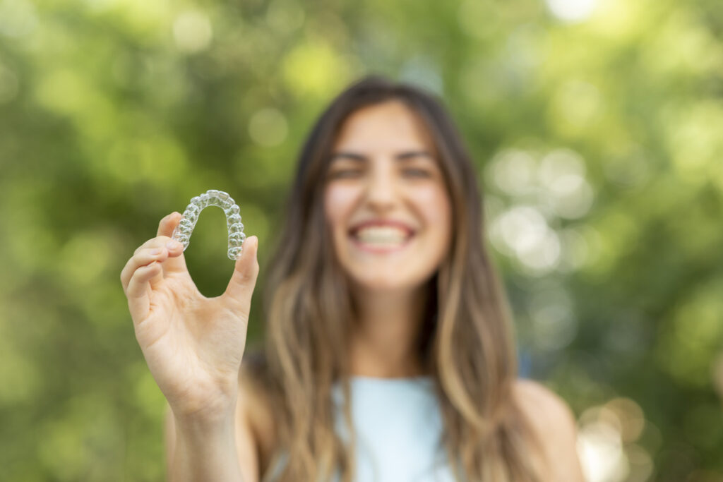 Close up of an Invisalign aligner being held by a smiling young woman on a warm summer day