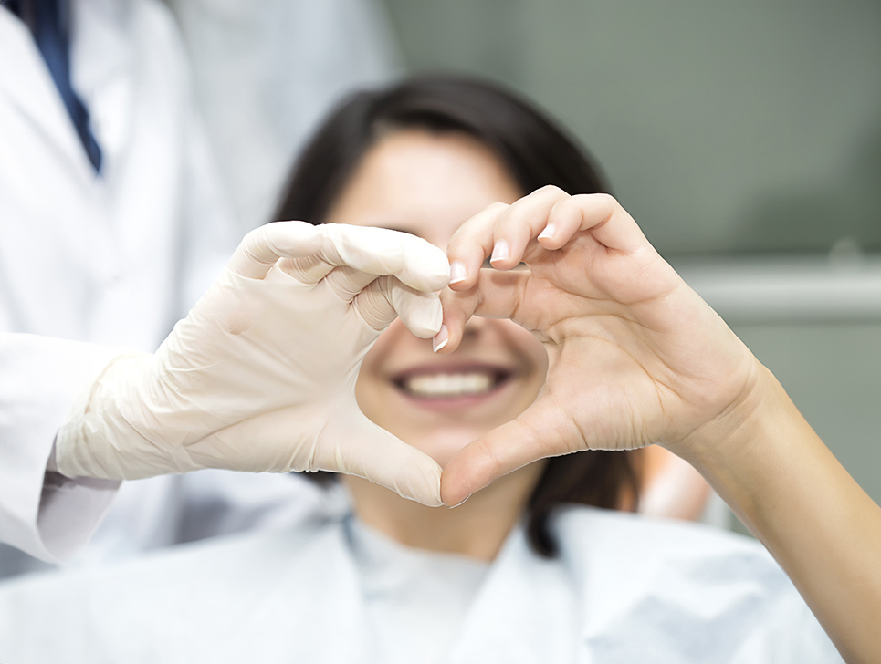 A smiling person sits in a dental chair, making a heart shape with their hands, while a gloved hand from a dental professional joins in.